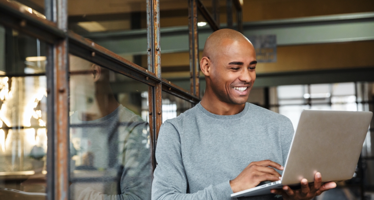 This images showcases a middle aged man leaning on the glass wall having a virtual meeting. He is holding a laptop in his hands and is smiling