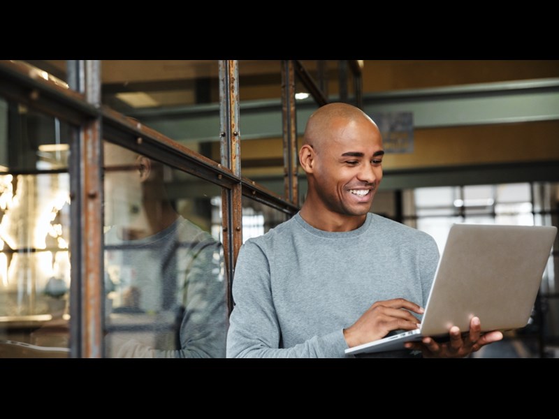 This images showcases a middle aged man leaning on the glass wall having a virtual meeting. He is holding a laptop in his hands and is smiling