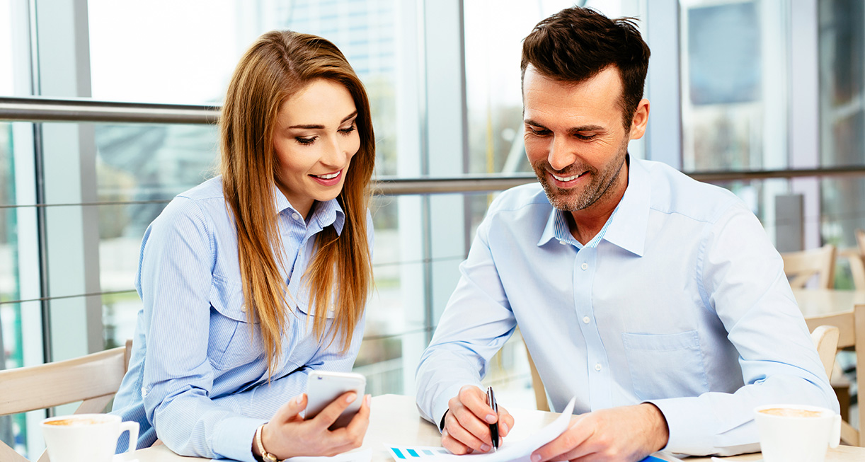 A man pointing to his paper with a pen and a lady next to him smiling with a phone in her hand